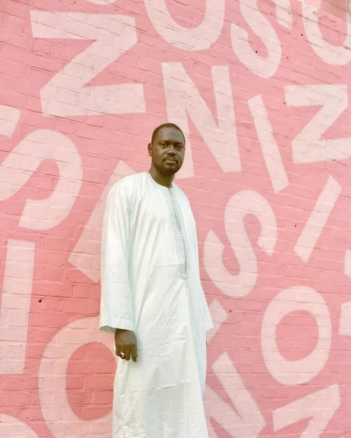 Portrait of an adult male in a light blue robe standing in front of a pink brick wall with white lettering for fashion content.
