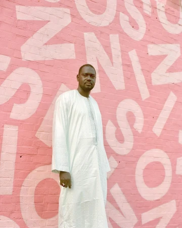 Portrait of an adult male in a light blue robe standing in front of a pink brick wall with white lettering for fashion content.