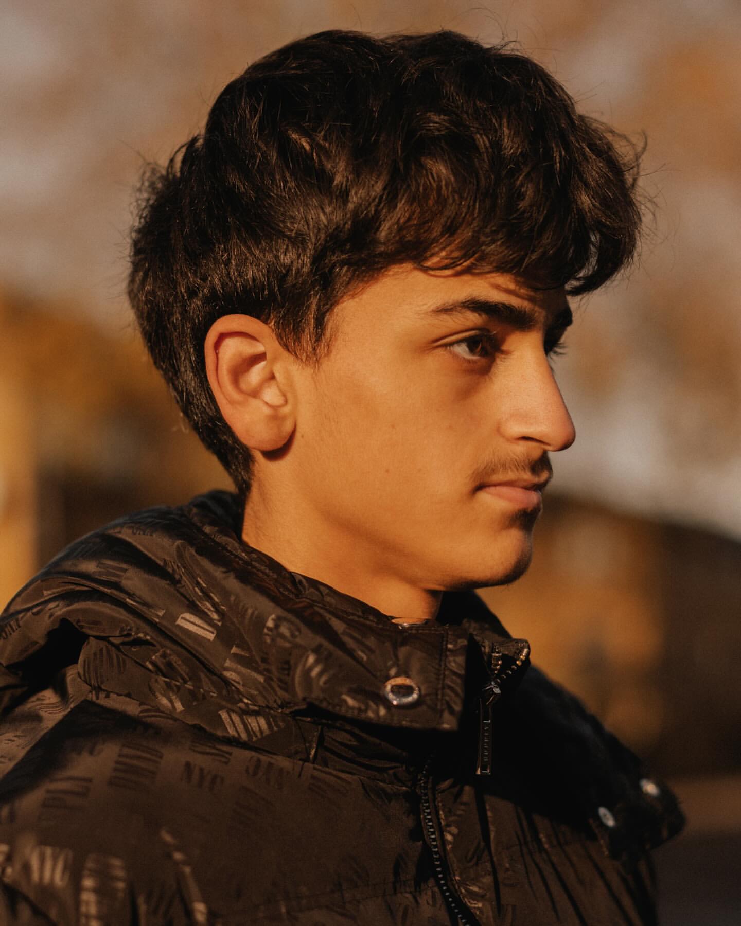 Portrait of a young adult male with dark hair and a slight mustache, wearing a black puffer jacket, set against a blurred background.