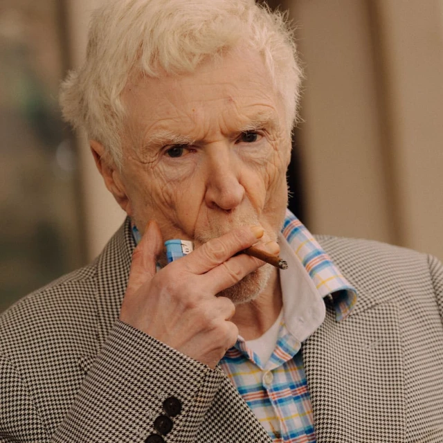 Older adult male with white hair in houndstooth blazer, smoking a cigar, close-up shot, indoor setting.