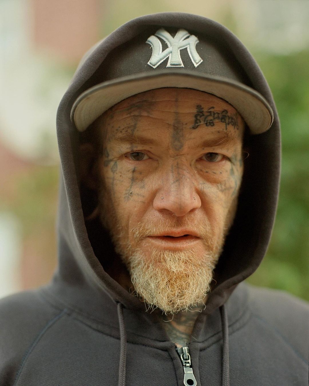 Close-up portrait of a man with face tattoos and gray beard wearing a baseball cap and hooded sweatshirt, outdoors, eye-level angle.