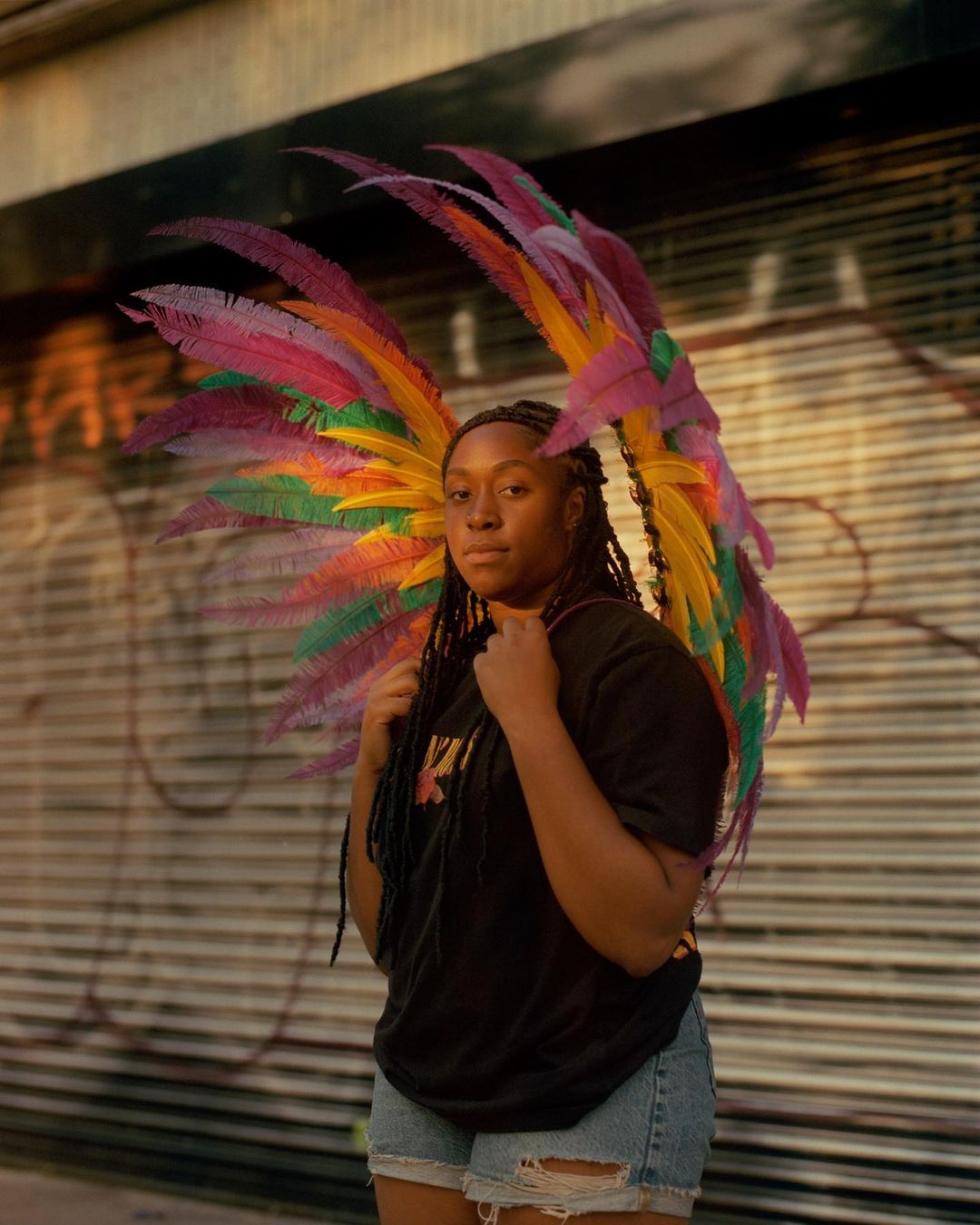 Portrait of a young Black woman with dark hair wearing a vibrant feather headdress, t-shirt and jean shorts standing against an urban background.