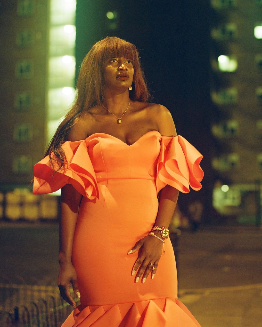 Black woman in coral off-the-shoulder dress poses against a dark building at night, accessorized with a watch and jewelry.