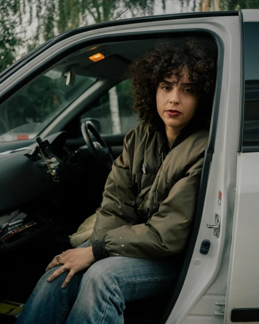 Portrait of a young woman with curly hair sitting in a car wearing a bomber jacket and jeans, looking at the camera with a neutral expression.