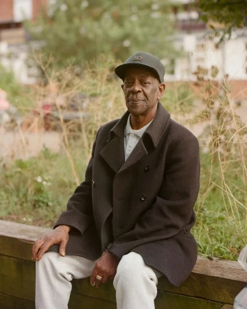 Elderly Black man in dark baseball cap and dark coat seated outdoors in London