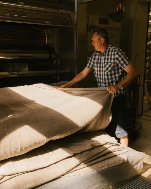 Man working with textiles in industrial setting, arranging folded cloths near machinery and light, dressed in checkered shirt.