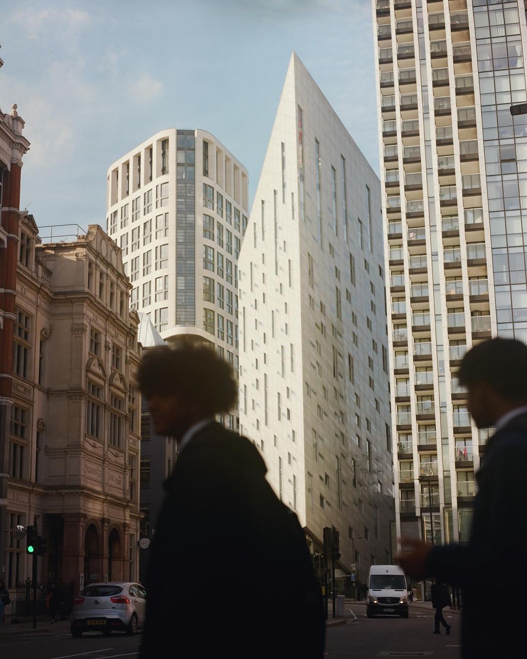 Urban scene featuring two silhouetted figures walking along a city street with modern buildings and a white van in the background.