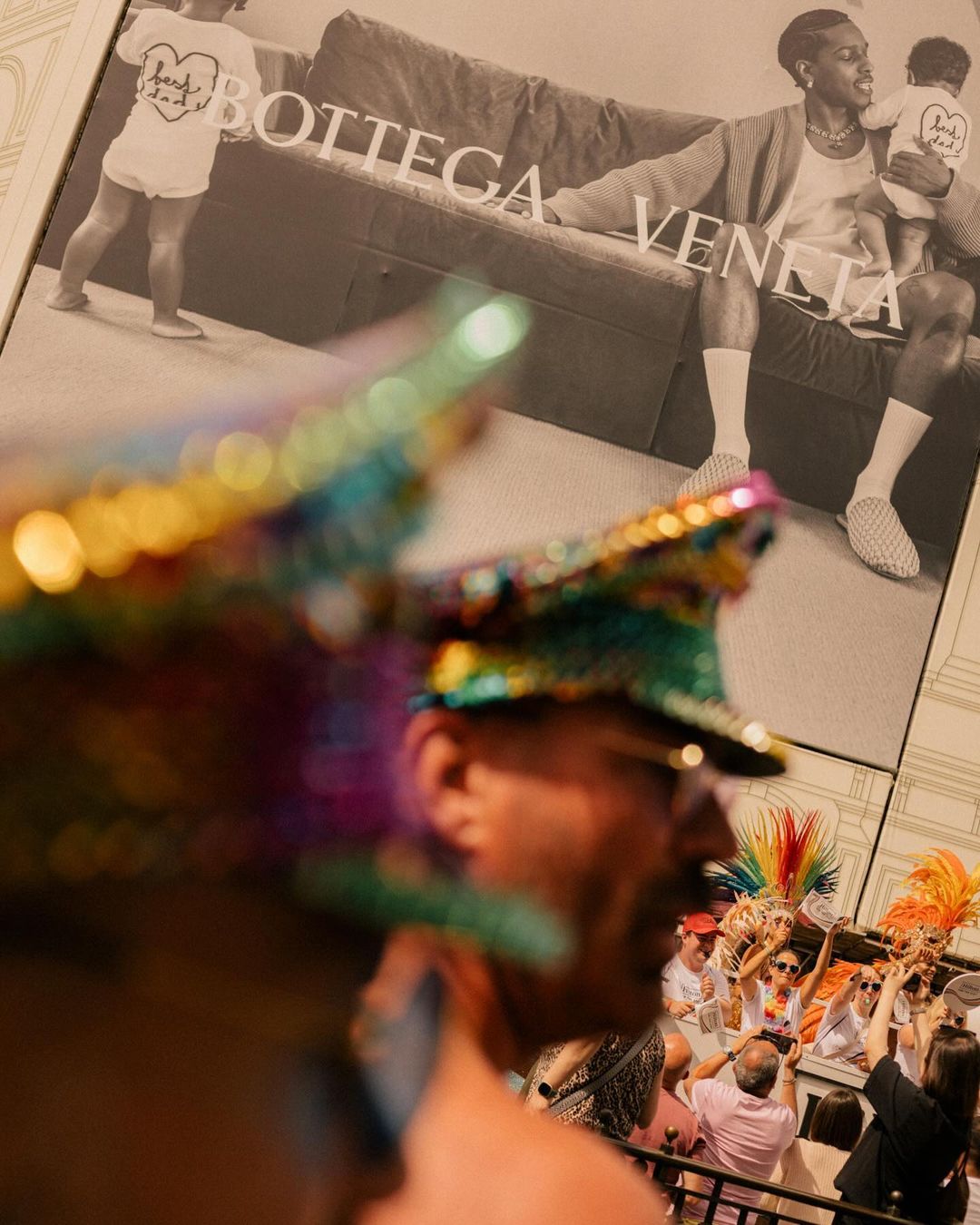 Pride parade crowd with a man in sequined hat foreground, ad with Black man holding child saying Bottega Veneta background.