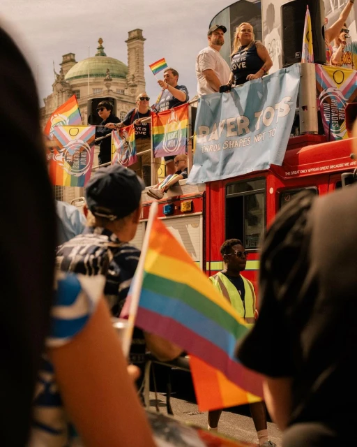 Pride parade features people on a decorated double-decker bus, holding rainbow flags with buildings in the background on a sunny day.
