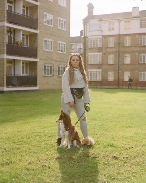 Adult woman stands with two dogs on a grassy lawn in front of a brick apartment building in the city on a sunny day.
