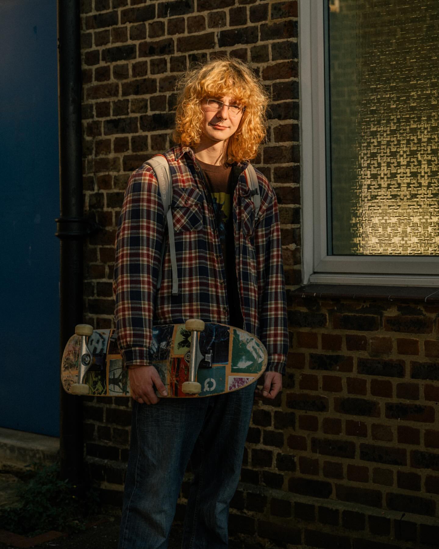 Young man with curly blonde hair wearing a plaid shirt holding a skateboard outside