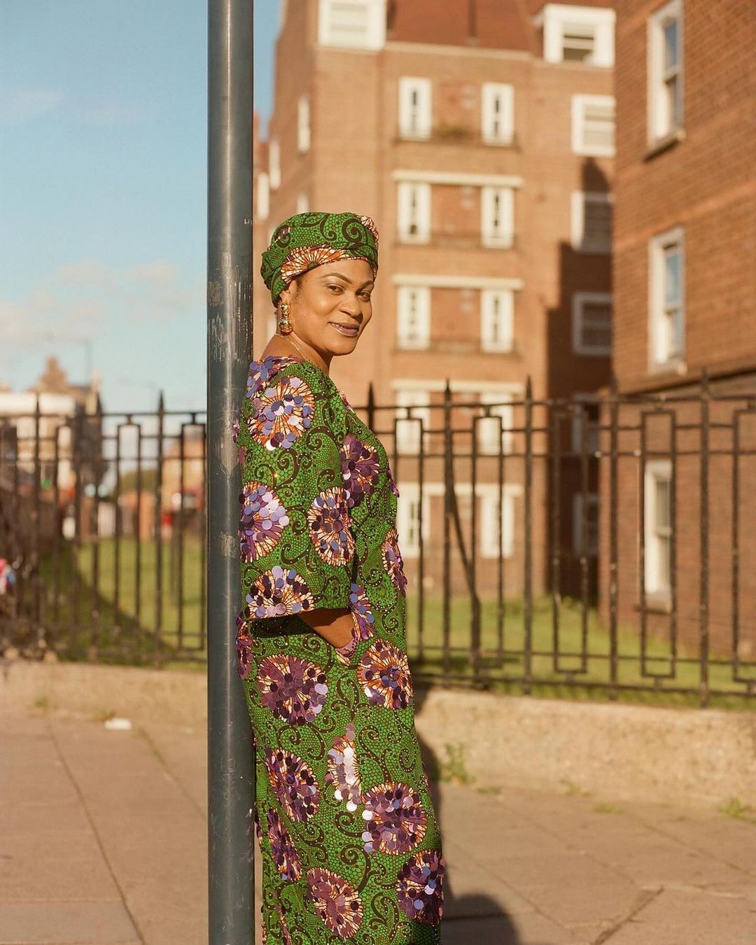 Woman in green floral dress and headwrap poses outdoors by a metal pole, with brick buildings in the background on a sunny day.