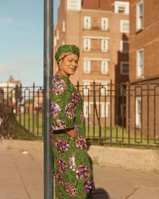 Woman in green floral dress and headwrap poses outdoors by a metal pole, with brick buildings in the background on a sunny day.