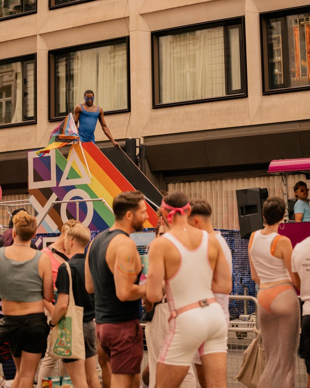 Pride parade scene with a Black male in blue tank top standing on a rainbow-themed float, with a crowd and a tall building behind him.