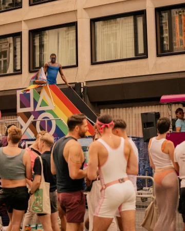 Pride parade scene with a Black male in blue tank top standing on a rainbow-themed float, with a crowd and a tall building behind him.