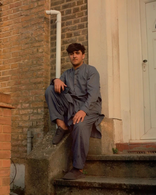 Portrait of a young adult man sitting on concrete stairs, dressed in a traditional grey salwar kameez, with a brick wall backdrop.