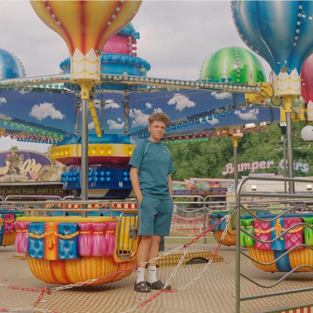 Young man in teal casual outfit stands on colorful amusement ride at an outdoor fair or carnival on a cloudy day; medium shot.