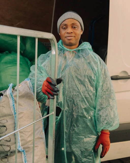 Man in a clear plastic raincoat and red gloves stands in front of a white van, holding onto a metal cart filled with green bags.