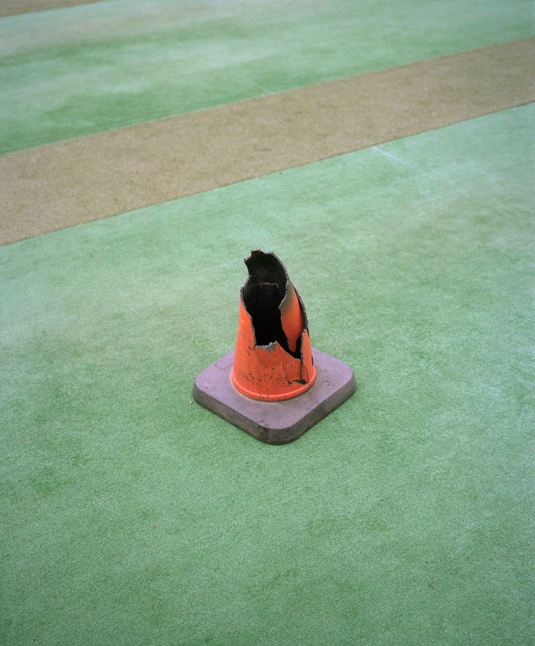 Damaged orange and gray traffic cone sits on green artificial turf with tan and white stripes in the background, outdoors.
