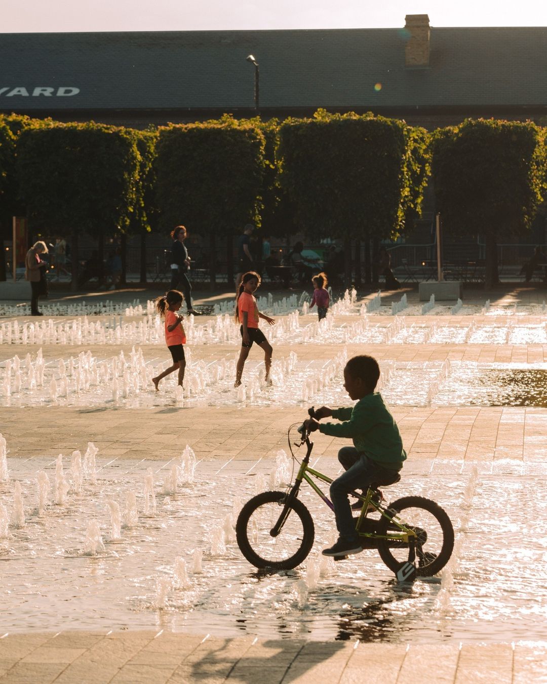 Boy riding bicycle through water fountain in urban park, several children playing in background, building with text on top, trees.