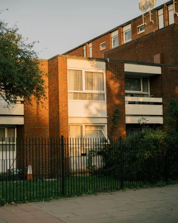 Exterior shot of a red brick apartment building with white-framed windows, balconies, black iron fence, and green foliage in an urban setting.