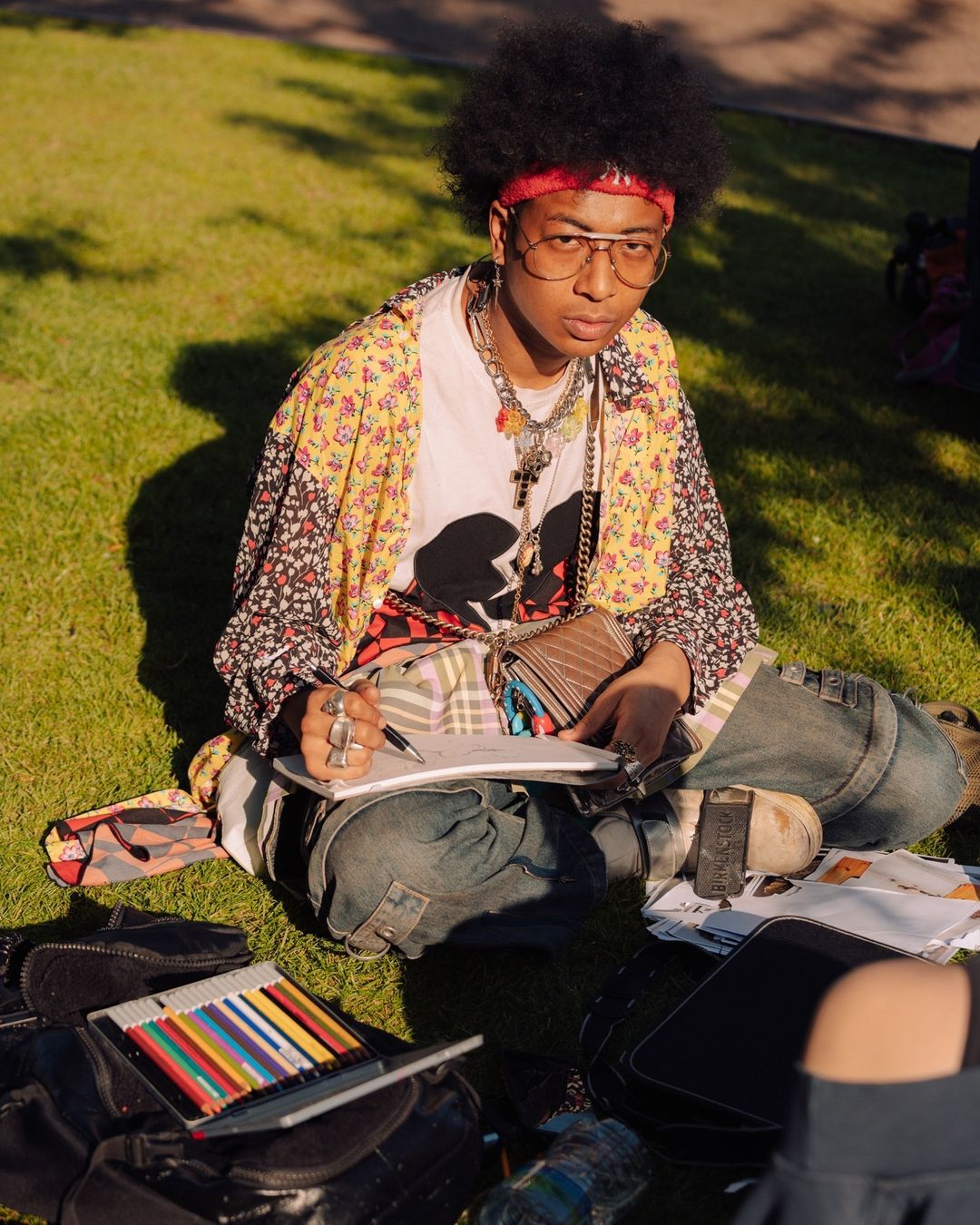 Young adult sitting cross-legged on grass, drawing in a sketchbook. Wearing a patterned jacket, jewelry, and glasses in outdoor light.