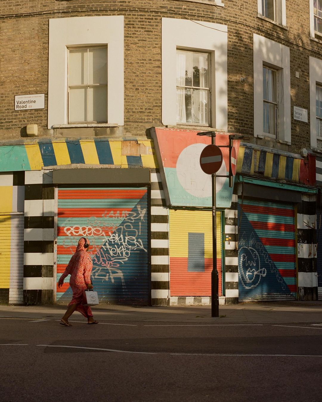 Pedestrian in patterned jumpsuit walks past colorful painted storefront shutters on a city street in London on a sunny day.