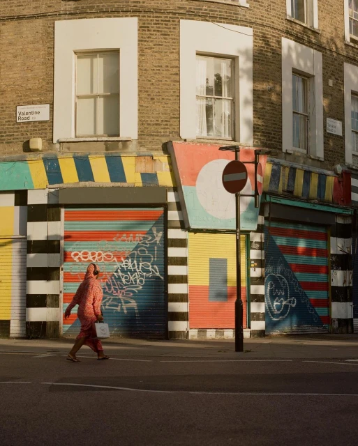 Pedestrian in patterned jumpsuit walks past colorful painted storefront shutters on a city street in London on a sunny day.