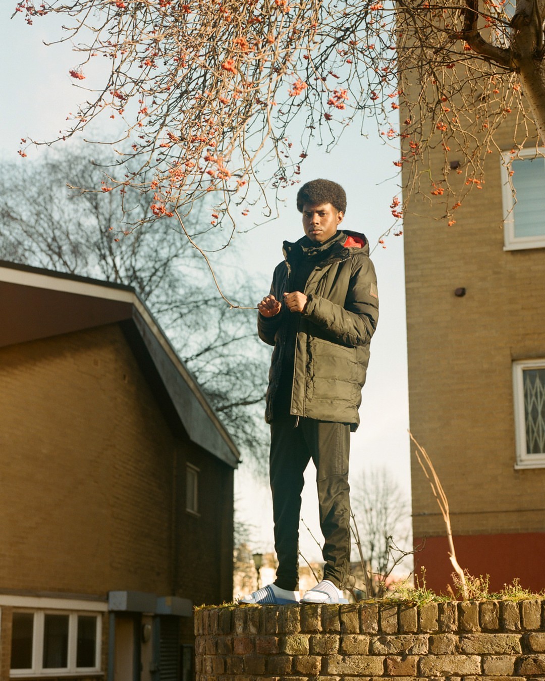 A young Black man in a puffy jacket standing on a brick wall in front of buildings and bare tree branches under a blue sky.