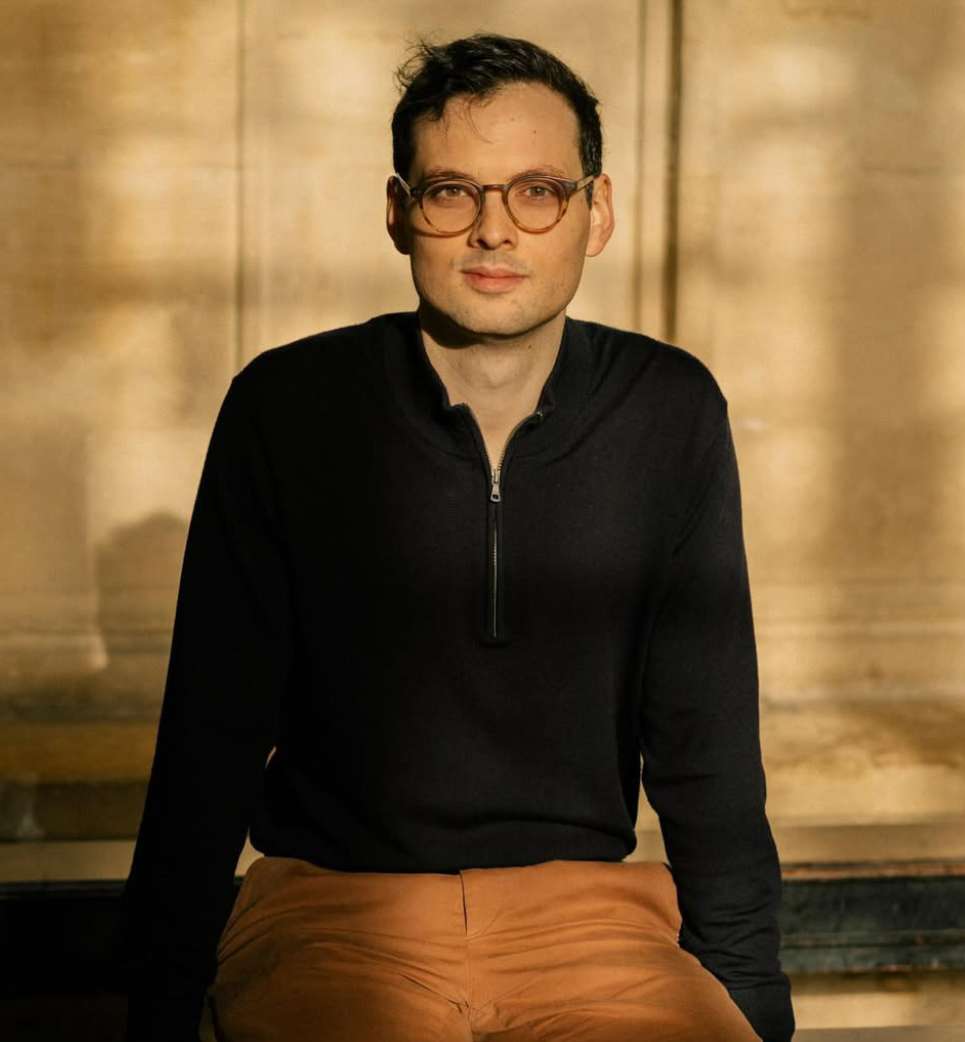 Portrait of a man wearing round glasses, black zip-up top, and brown trousers outdoors by Freya Najade