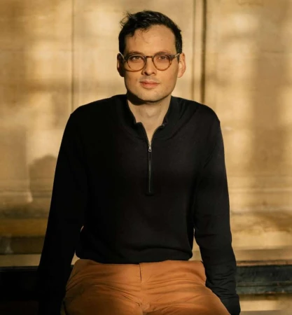 Portrait of a man wearing round glasses, black zip-up top, and brown trousers outdoors by Freya Najade