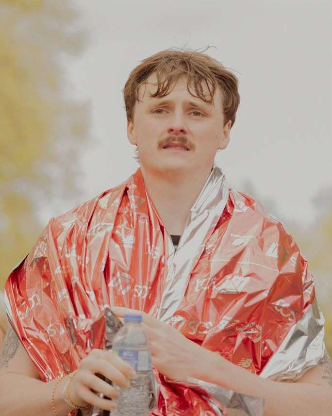 Portrait of an adult male wrapped in a red and silver foil blanket, holding a clear plastic water bottle, outdoors during daytime.