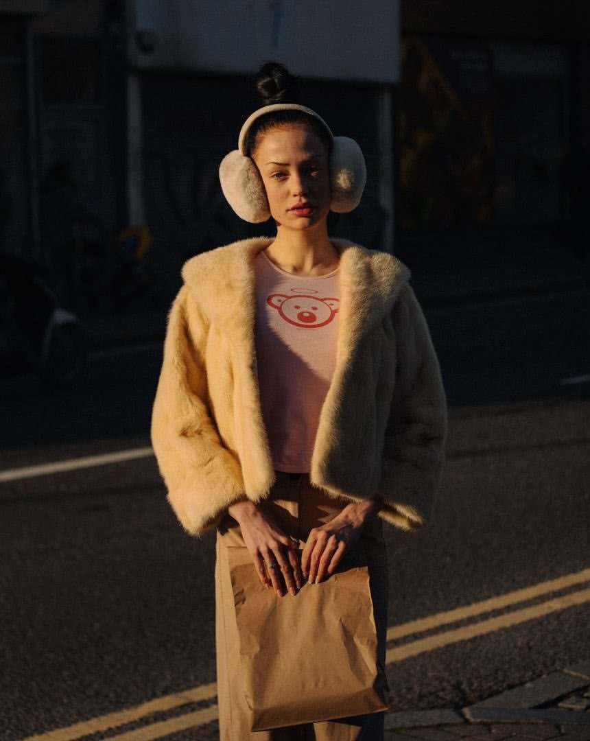 Woman in fur coat and earmuffs standing on street. A paper bag is in her hands. Faded building visible in the background.