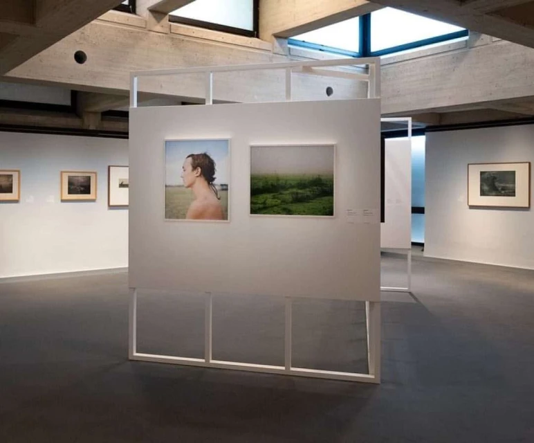 Art gallery exhibit featuring two framed images, one of a man in profile and one of a green field, with white walls and concrete architecture.