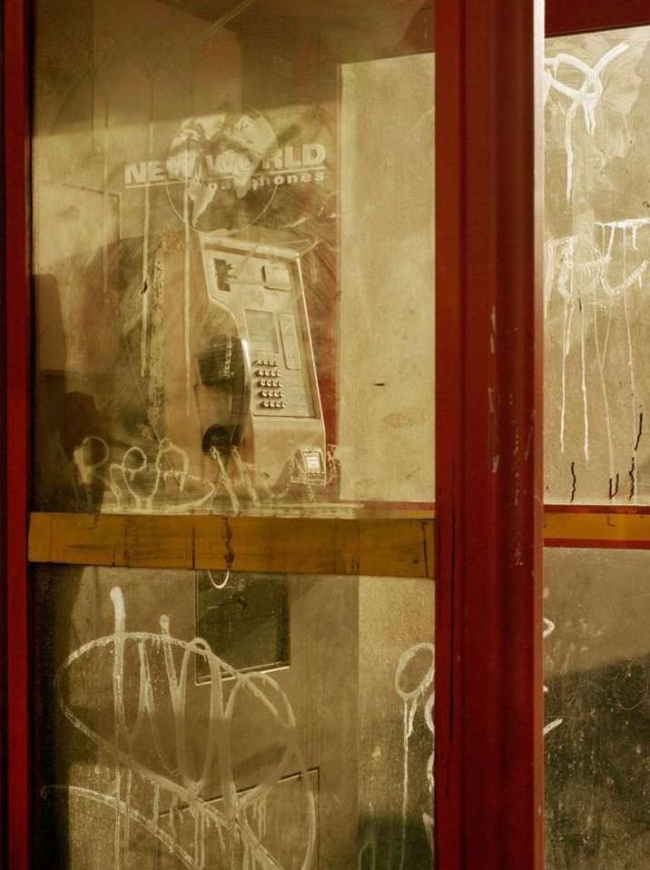 An old, graffiti-covered phone booth with peeling paint stands on a city street at night, casting long shadows under dim lighting.