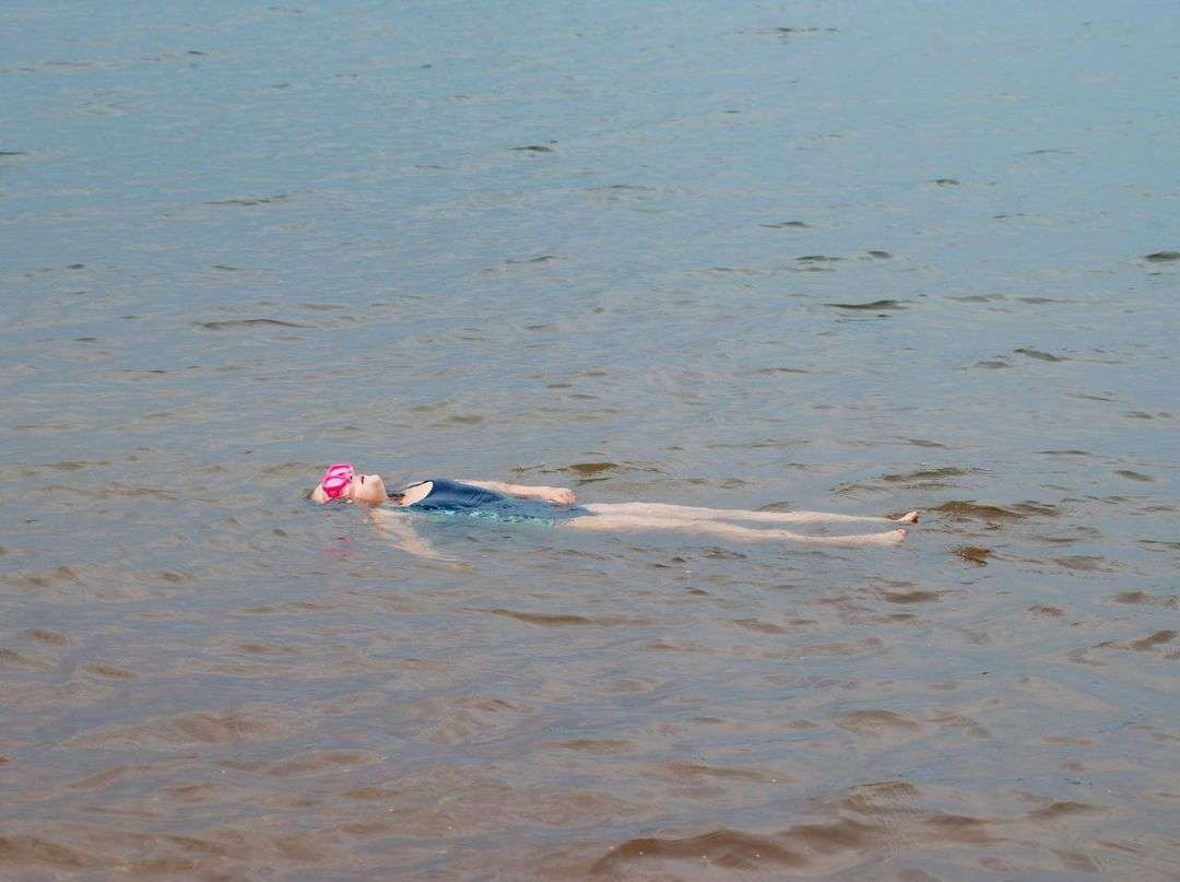 A young woman in a blue swimsuit and pink swim cap floats on her back in a calm lake or ocean under soft natural light.