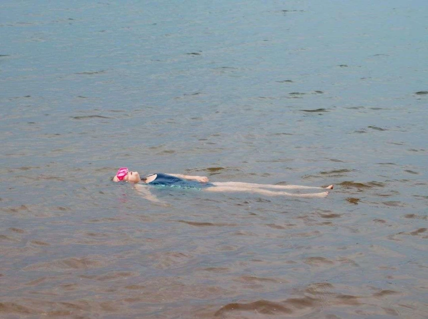 A young woman in a blue swimsuit and pink swim cap floats on her back in a calm lake or ocean under soft natural light.