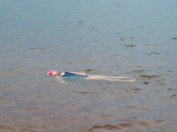 A young woman in a blue swimsuit and pink swim cap floats on her back in a calm lake or ocean under soft natural light.