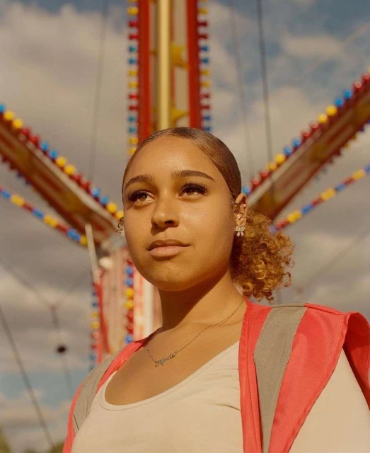Portrait of a young woman wearing a reflective vest looking up, with a colorful amusement park ride in the background against a cloudy sky.