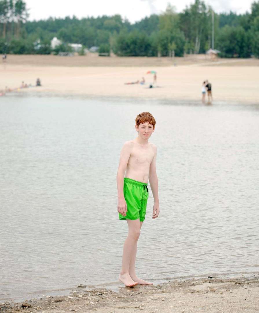 A fair-skinned child with red hair stands in green swim trunks on the edge of a sandy beach and clear water on a bright day.