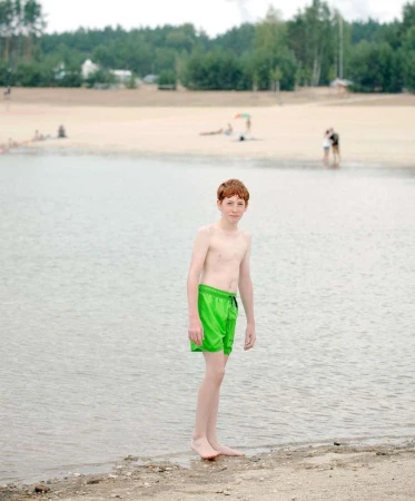 A fair-skinned child with red hair stands in green swim trunks on the edge of a sandy beach and clear water on a bright day.