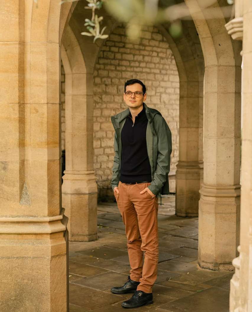 An adult man with glasses stands with hands in pockets in an archway with stone pillars and walls, wearing an olive jacket and brown pants.