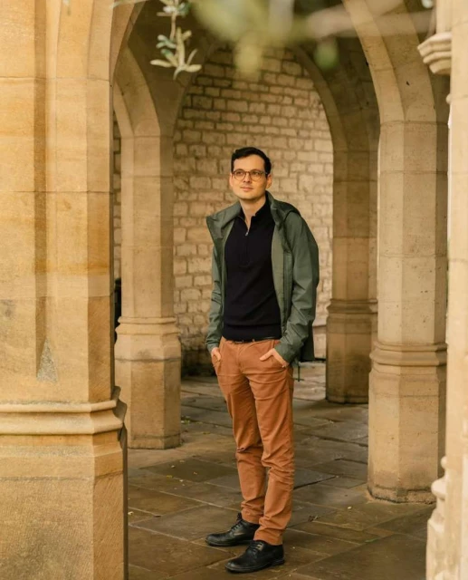 An adult man with glasses stands with hands in pockets in an archway with stone pillars and walls, wearing an olive jacket and brown pants.