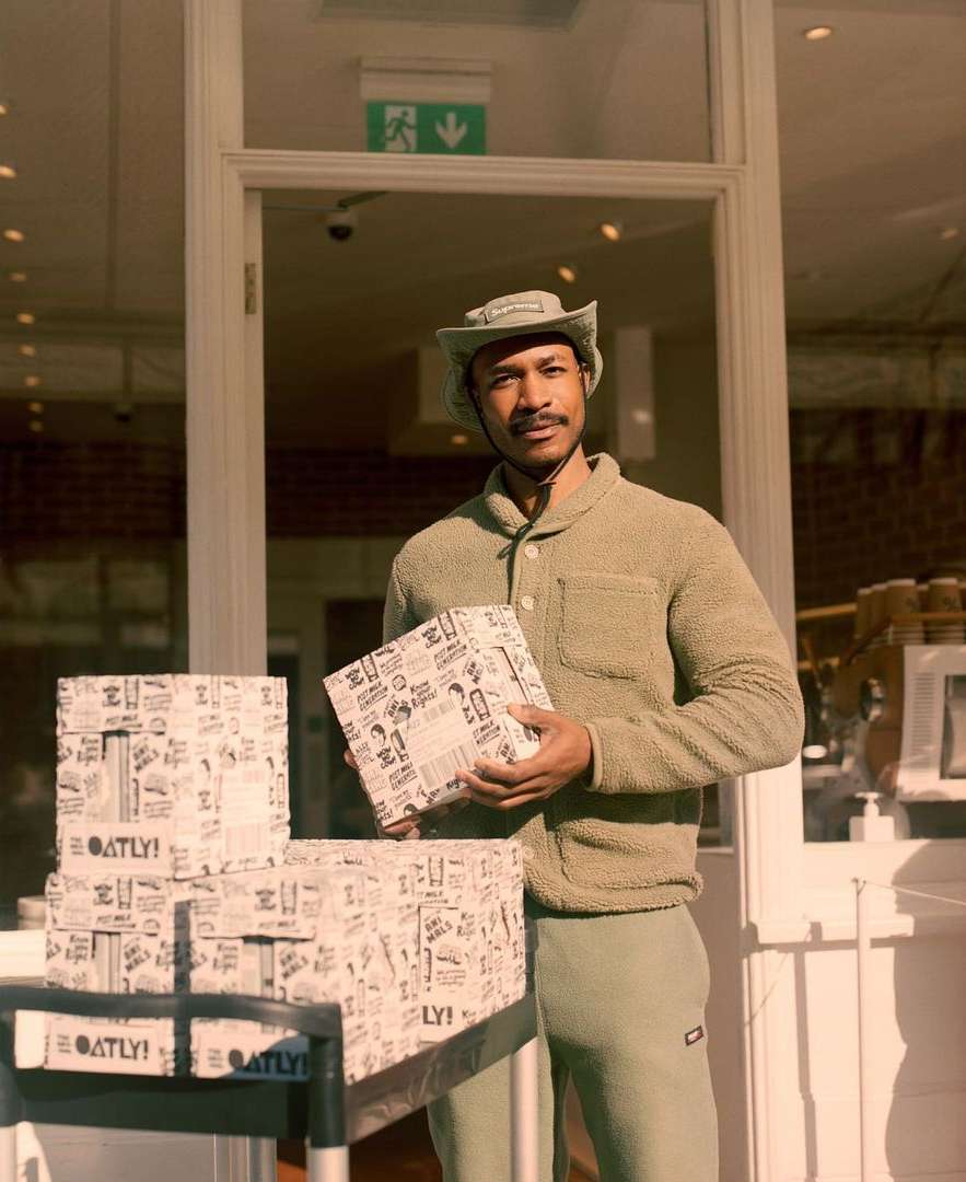A man in a hat and jacket standing outside a shop with boxes of Oatly product, posing in front of a white-framed glass doorway.
