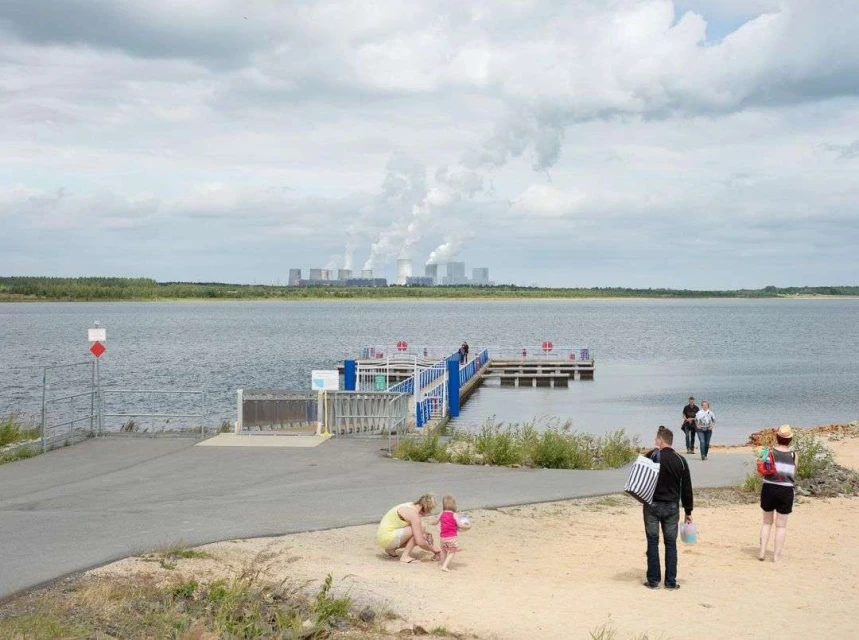 People enjoy a day at the lakeside with a pier and distant industrial buildings under a cloudy sky, creating a recreational scene.