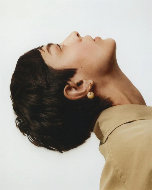 Adult woman poses, head tilted back, wearing gold hoop earring and tan collared shirt against white seamless backdrop in studio setting.