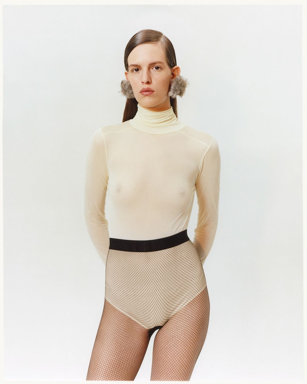 Woman in a turtleneck top, textured brief, and furry earrings poses in a studio with neutral lighting and a white background.