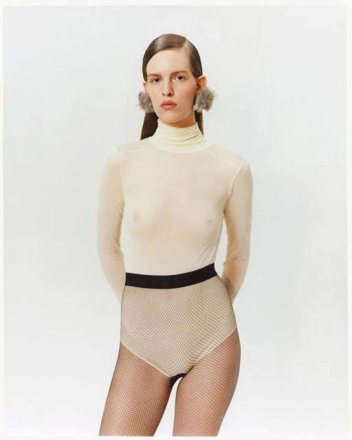 Woman in a turtleneck top, textured brief, and furry earrings poses in a studio with neutral lighting and a white background.
