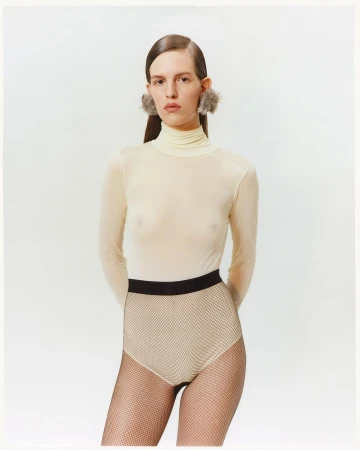 Woman in a turtleneck top, textured brief, and furry earrings poses in a studio with neutral lighting and a white background.