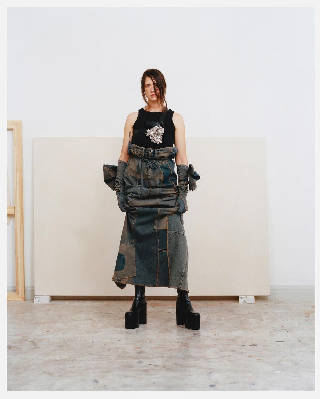 A slender adult woman stands in a photo studio, wearing a dark top, a dark denim patchwork skirt, platform boots, and matching gloves.